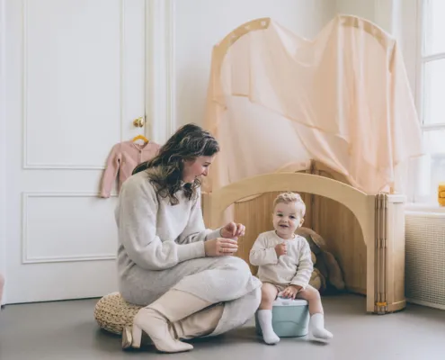 Mother sitting on the floor with a baby on a small potty in a bright, pastel nursery, both smiling.