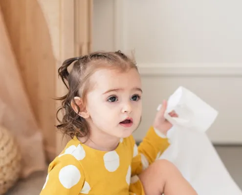 Young toddler in a yellow polka-dot dress sits on the floor, holding a tissue and looking off to the side in a bright room.