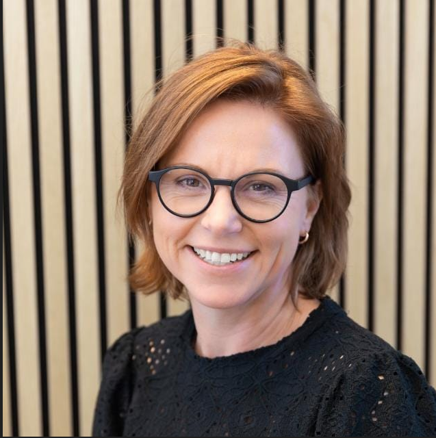 Portrait of a smiling woman with short brown hair and round black glasses, wearing a black lace top, against a striped background.